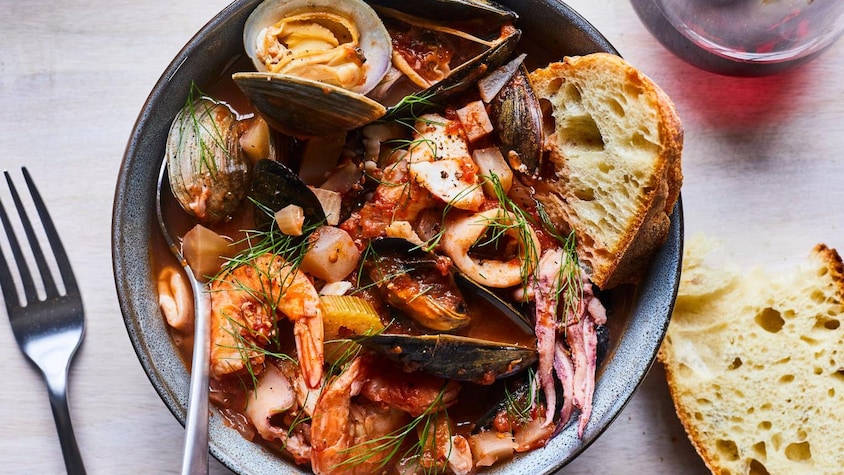 Une soupe de fruits de mer dans un bol accompagnée d'un morceau de pain et d'une cuillère, près d'une fourchette et d'un bout de pain.