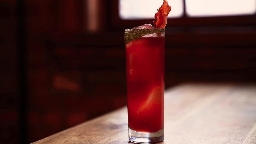 Un cocktail rouge dans un verre allongé avec une tranche de bacon en décoration.