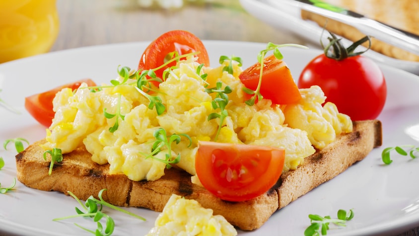 Des œufs brouillés sur une toast. Le tout est garni de morceaux de tomate et d'herbes fraîches.