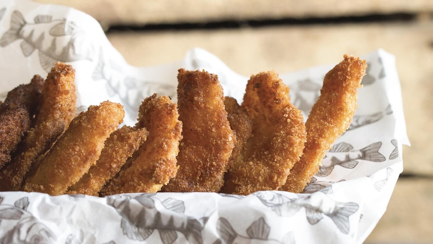 Des filets de poissons panés sont placés en dominos dans un papier craft, dans un plat rectangulaire en bois.