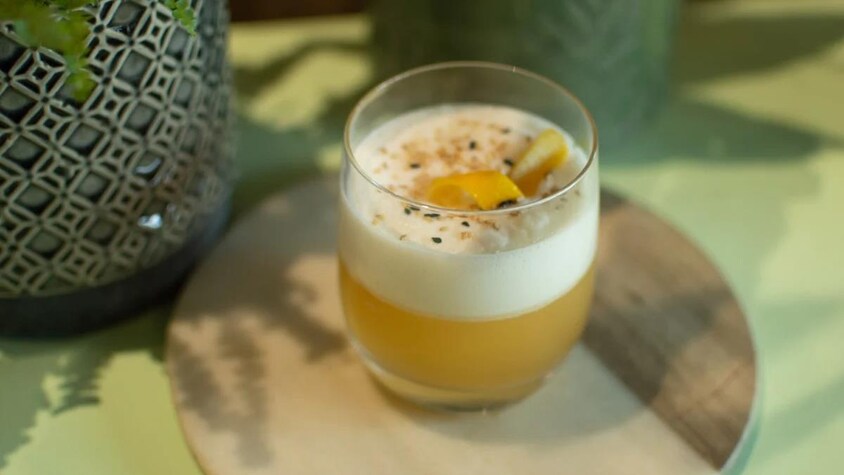 Un verre contenant un mocktail Hojicha sour avec sa garniture de zeste de citron et graines de sésames.