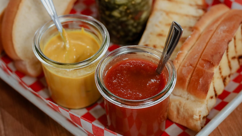 Des pots de moutarde et de ketchup servis dans un cabaret, aux côtés de pains à hot-dog et de relish.