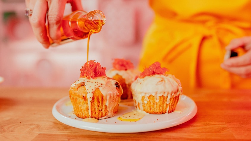 Une main qui verse du sirop de safran sur 3 muffins décorés avec du glaçage blanc et des carottes râpées.