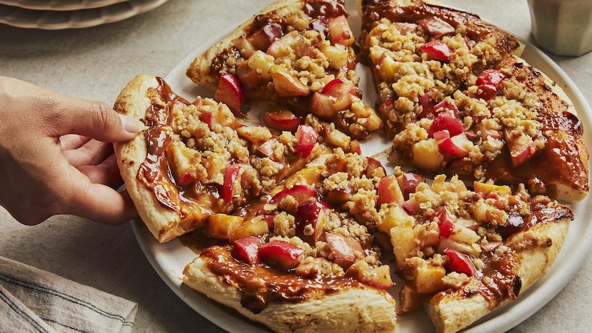 Dans une grande assiette, une main prend une pointe de la pizza dessert aux pommes. Autour, il y a une pile d’assiette et une petite tasse de café.