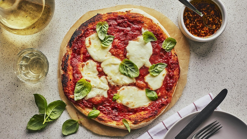 Au centre d’une table, une pizza Margherita est posée sur un papier parchemin rond. Autour, des feuille de basilic frais, une bouteille de vin blanc, un petit pot d’huile épicée et une assiette.