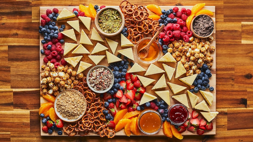 Une planche de bois est garnie de morceaux de gâteau au fromage, de bleuets, de framboises, de fraises, de pretzels, de tranches d'oranges, de pacanes, de pistaches, de chocolat, de confiture, de miel, de Cracker Jack et de flocons d'érable.