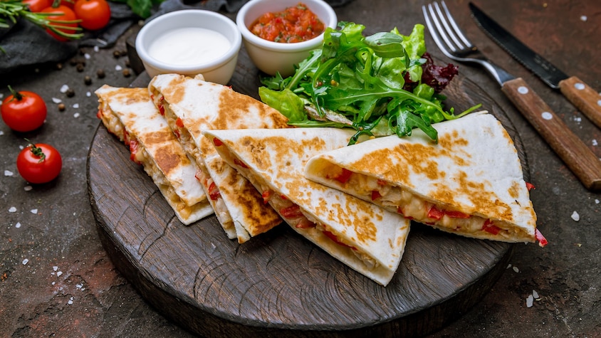 Quatre morceaux de quesadilla au poulet dans une assiette avec un peu de salade, un petit pot de salsa et un petit pot de crème sure.