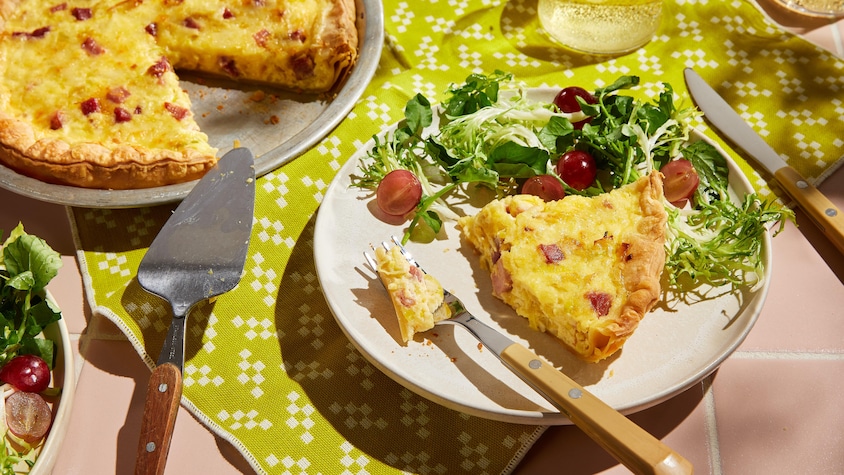 Une portion de quiche au jambon dans une assiette. Elle est servie avec une salade verte et des raisins frais.