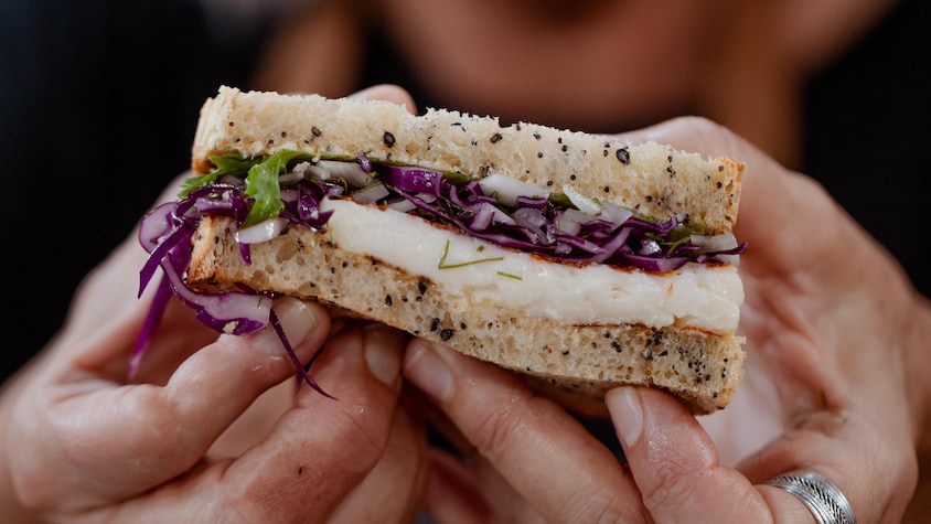 Une part de sandwich au fromage, salade de chou rouge et fenouil dans les mains de la cheffe Josée Robitaille.