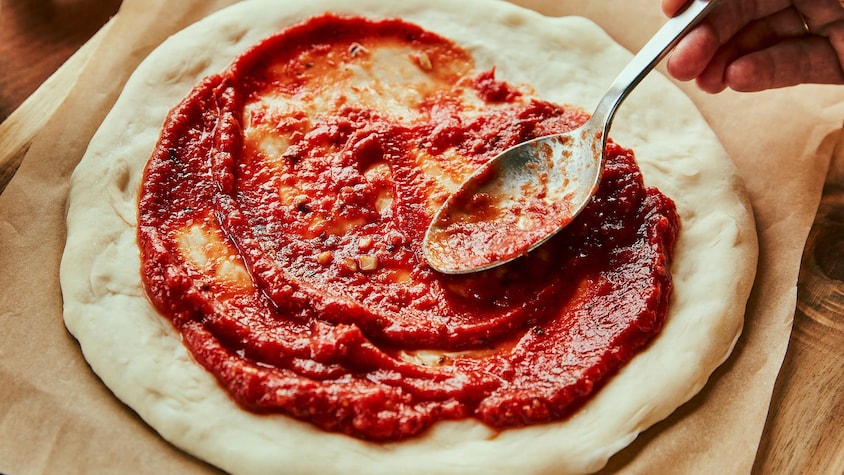 Une main étend la sauce à pizza sur une pâte à pizza non cuite avec une cuillère. 