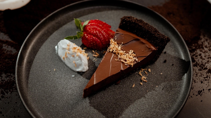 Un morceau de tarte au chocolat dans une assiette avec un peu de crème fouettée à la noix de coco et une fraise tranchée.