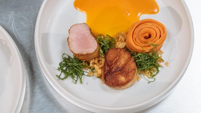 Dans une assiette se trouve un médaillon de filet de porc, un pétoncle grillé et une rosace de carottes. Le tout est arrosé d'une sauce orangée.