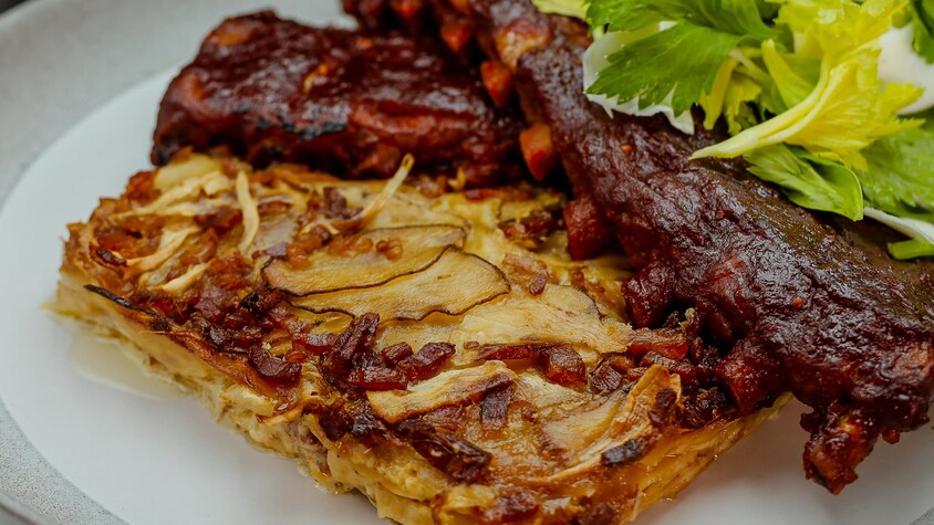 Un gratin de panais, pommes de terre rattes et bacon qui accompagne les côtes levées dans une assiette.