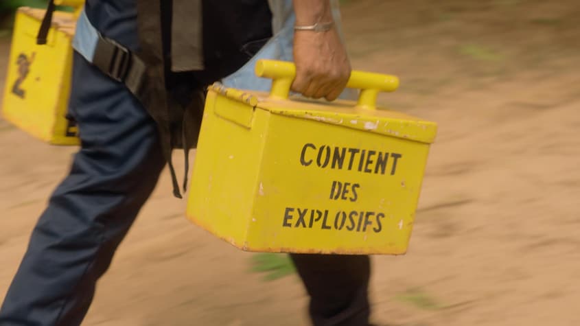 Une personne marche avec deux boîtes sur lesquelles ont peut lire Contient des explosifs.