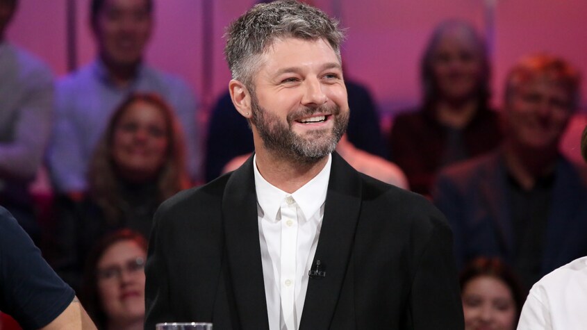 L'homme sourit. Sa barbe et ses cheveux sont grisonnants. Il porte une chemise blanche et un veston noir.