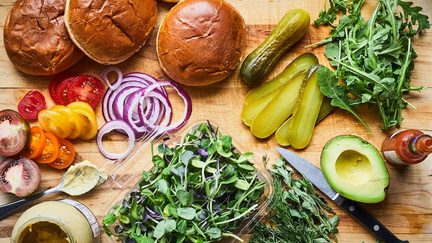 Sur un ilot de cuisine des pains à burgers ont été tranchés en deux, cinq tranches de cornichons, la moitié d'un avocat, des herbes fraîches et un pot de moutarde à Dijon y ont été aussi déposés. 