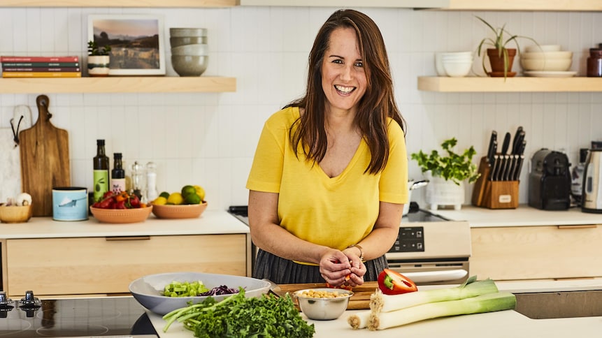 La nutritionniste Geneviève O'Gleman porte un chandail jaune et tient des légumes dans ses mains en souriant à la caméra. Plusieurs aliments sont déposés sur le comptoir de cuisine.