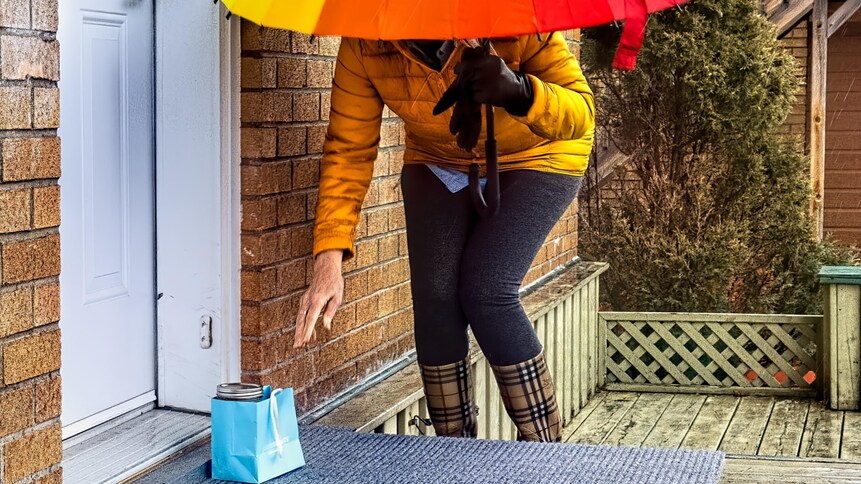 Devant le perron d'une maison,  à l'abri sous un parapluie, une personne est penchée pour prendre  un sac cadeau contenant de la nourriture.
