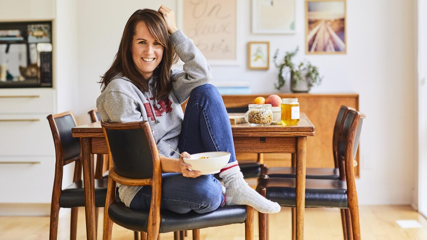 Geneviève O'Gleman assise sur une chaise de manière décontractée et tenant un bol à la main. Sur la table en arrière-plan, on voit du granola, du miel et des pommes.