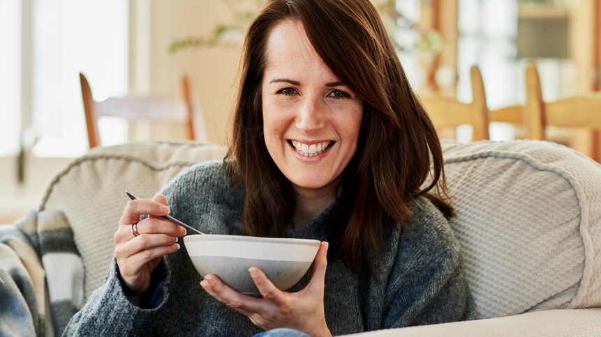 Geneviève O'Gleman assise sur un canapé et qui tient un bol de soupe.