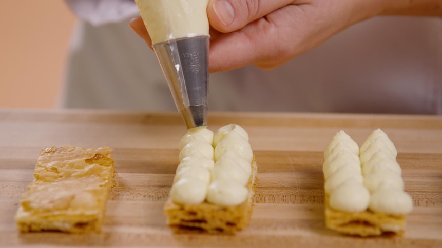 Une personne garnit de crème pâtissière un rectangle de pâte feuilletée. 