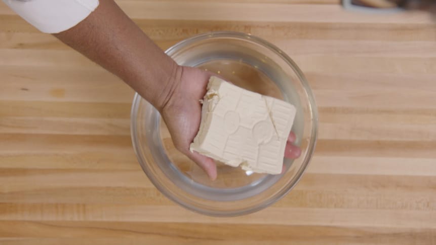 Une personne tient un bloc tofu fait maison. Il égoutte celui-ci au-dessus d'un bol en verre. 