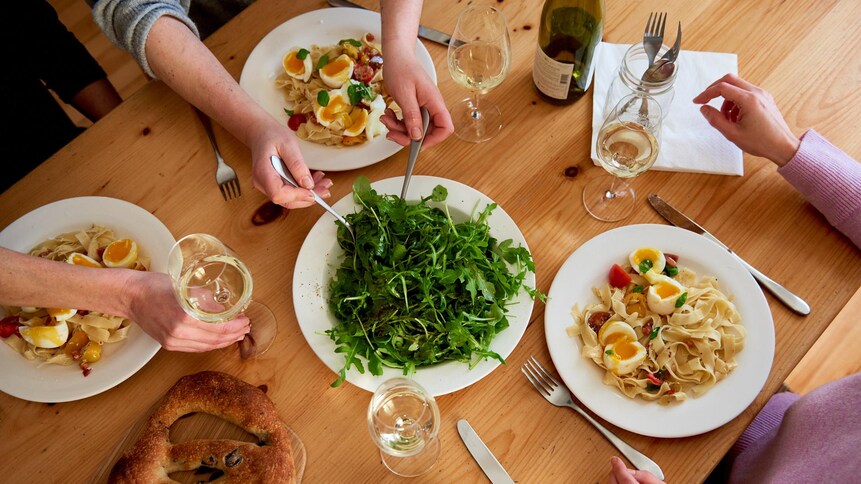 Une tablée garnie d'assiettes de pappardelles aux oeufs mollets, d'une salade de roquette, d'une fougasse aux olives et de verres de vin. Les convives s'apprêtent à commencer à manger.