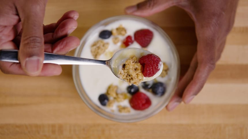 Une personne tient dans sa main une petite cuillère. Celle-ci contient du yogourt, un morceau de granola et une framboise mûre. 