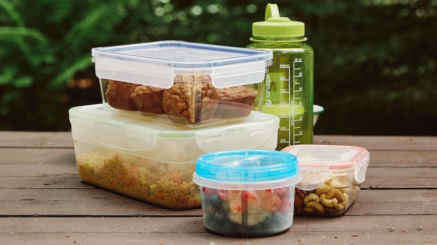 Sur une table à pique-nique, est déposé plusieurs contenants en plastique qui eux, contiennent une salade de couscous, des petits fruits et des noix. Il y a aussi une bouteille d'eau sur la table. 