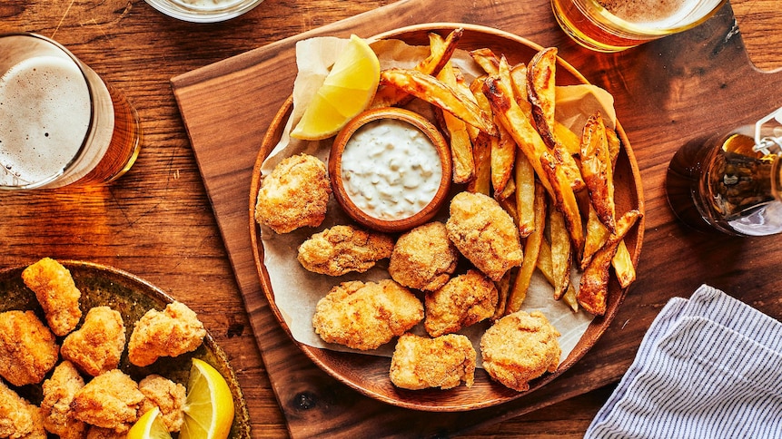 Des bouchées de fish and chips dans une assiette en bois sur une planche de bois, accompagnés de frites, d’une sauce tartare et de verres de bière. 