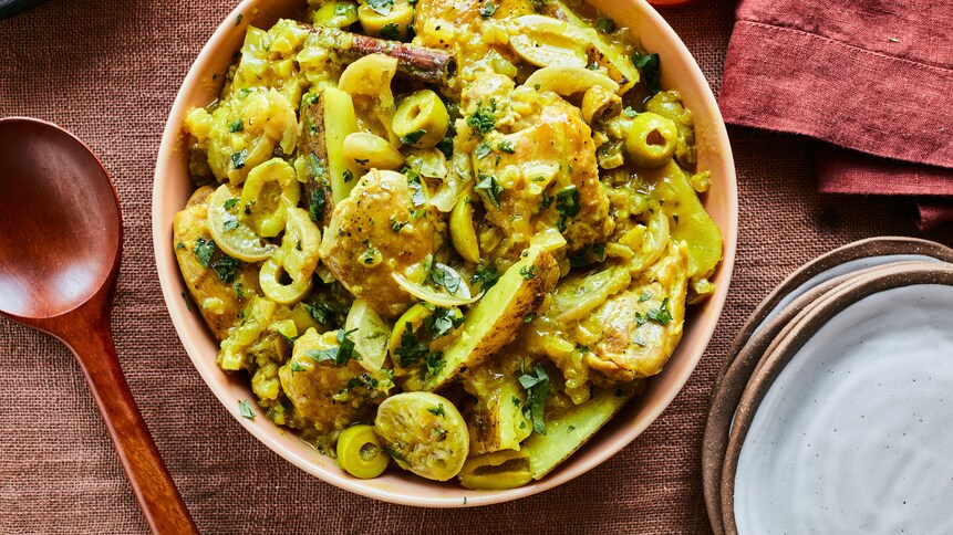 Il est possible de voir un bol contenant le poulet au citron et aux olives à la marocaine au centre de la photo. Il y a une cuillère de bois à la gauche du bol, des assiettes et un linge de table à la droite. 