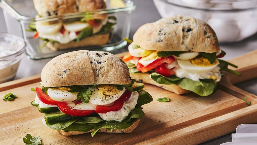 Trois sandwichs aux œufs à la grecque sur une planche de bois avec des plats pour les lunchs.