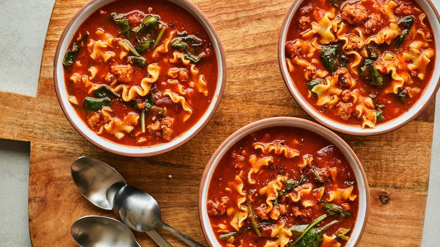 Sur une planche en bois est déposé trois bols et trois cuillères. Les bols sont remplis de soupe-lasagne. 