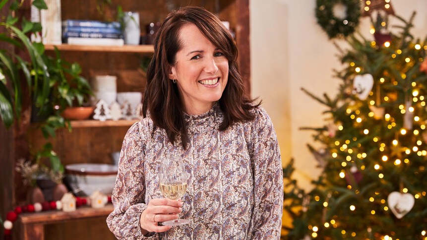 Geneviève O'Gleman debout devant un sapin de Noël avec un verre de vin à la main.