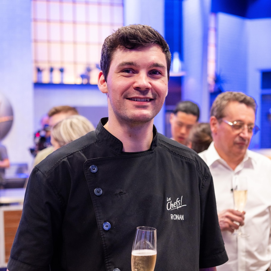 Le chef Ronan Ulliac dans la cuisine de l'émission Les chefs!, tenant une coupe de champagne dans la main droite.