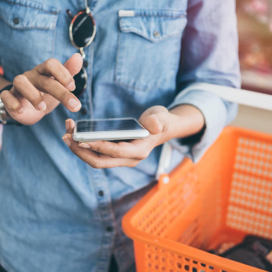 Une personne qui porte une chemise en denim tient un panier d'épicerie au creux du coude et un téléphone intelligent dans la main.