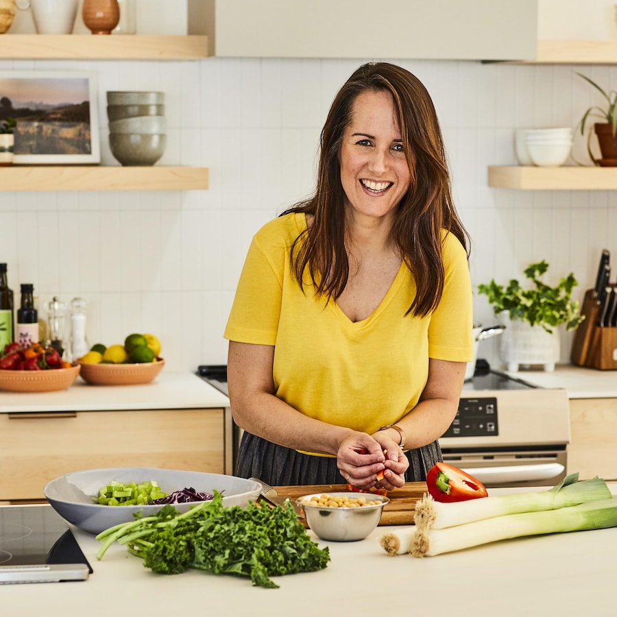 La nutritionniste Geneviève O'Gleman porte un chandail jaune et tient des légumes dans ses mains en souriant à la caméra. Plusieurs aliments sont déposés sur le comptoir de cuisine.