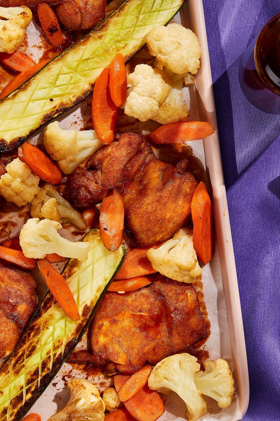 Des hauts de cuisses de poulet laqués de sauce BBQ, des tranches de carotte, des fleurons de chou-fleur et des courgettes sur une plaque à cuisson.