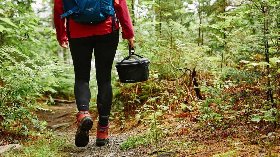 Une femme de dos en forêt qui transporte du matériel de camping.