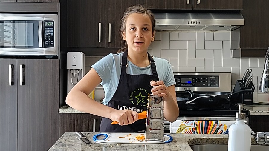 Une jeune fille en train de râper une carotte dans sa cuisine.
