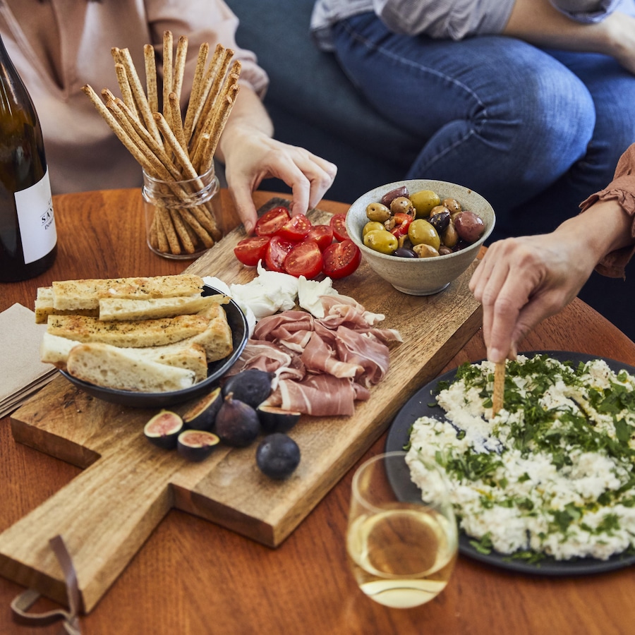 Les mains de quatre personnes autour d'une table de salon qui prennent l'apéro avec une bouteille et des verres de vin et une planche de bois avec charcuteries, pain, olives, figues, tomates ainsi qu'une assiette de fromage frais aux herbes.