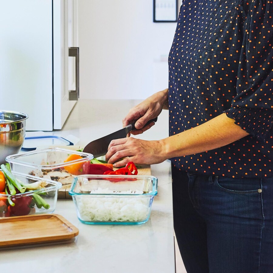 Une femme qui tranche des légumes sur un plan de travail avec différents plats réutilisables en verre.