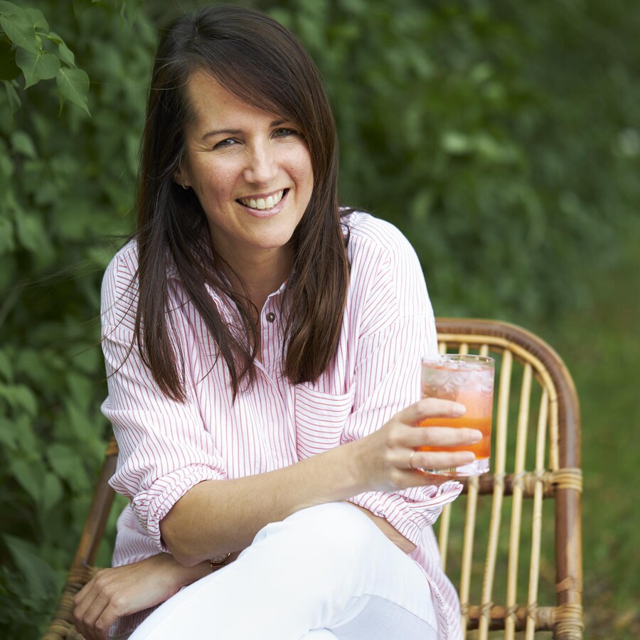 Geneviève O'Gleman assise sur une chaise en bois dans un jardin et tenant un cocktail dans sa main. 