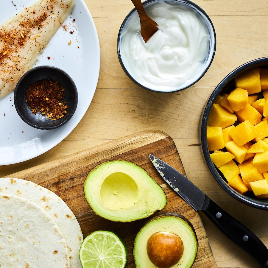 Un filet de poisson blanc cru dans une assiette avec des épices, des tortillas, un avocat, une lime et des cubes de mangue.