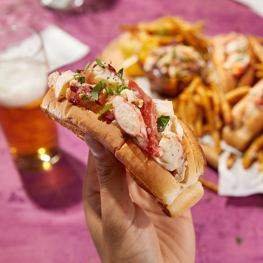 Une main tient un pain hot-dog farci de homard, en arrière-plan, un plateau de frites et de guédilles de homard. 