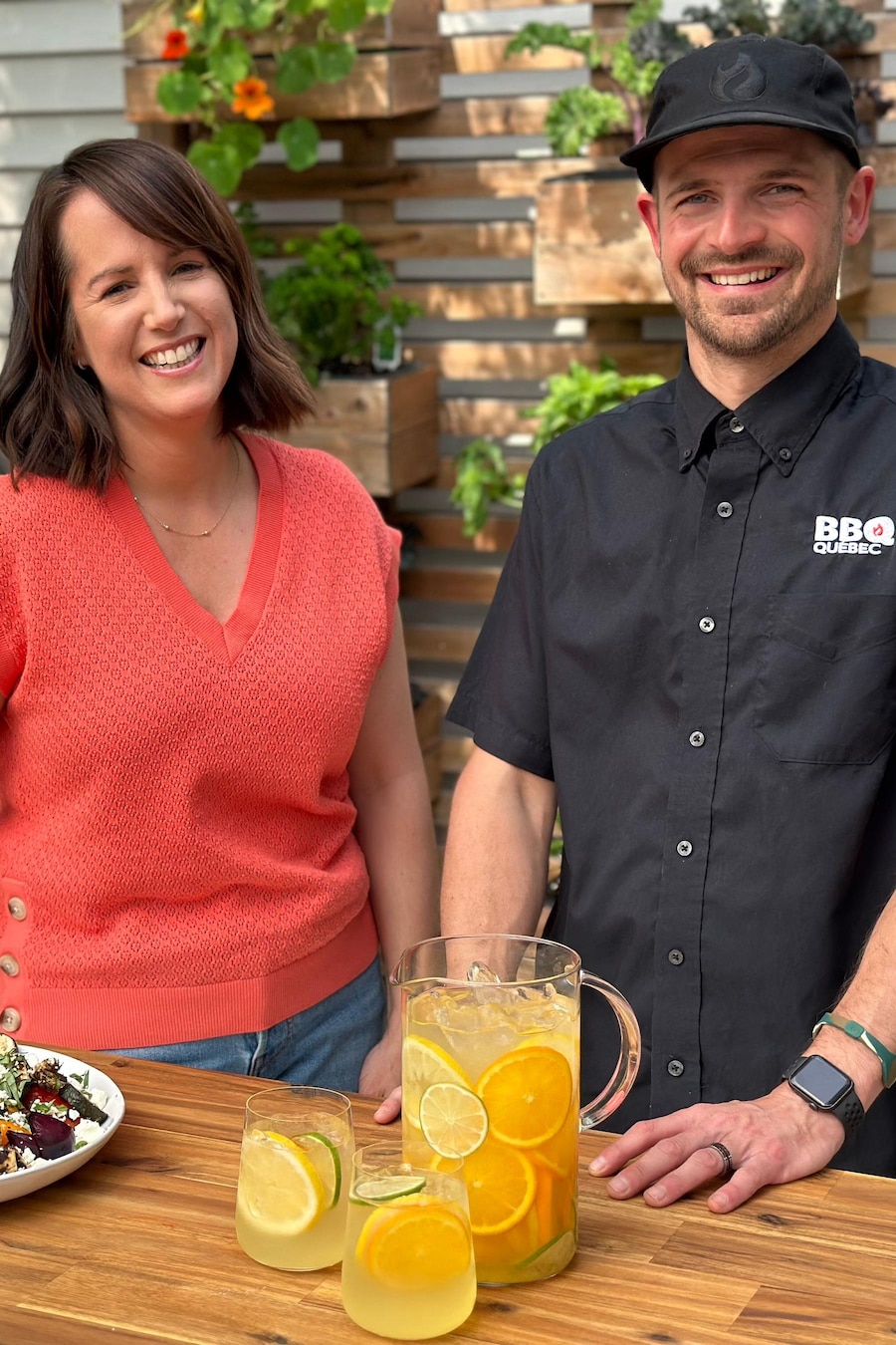 La nutritionniste Geneviève O’Gleman et le président de BBQ Québec, Max Lavoie, sont sur une terrasse à côté d’un barbecue et d’une table avec une agua fresca aux agrumes et un plat de poulet et légumes grillés à la grecque. 