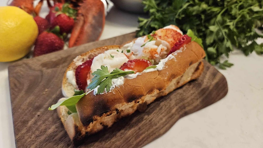 Un pain à hot-dog garni de homard, de tranches de fraises, d'herbes fraîches et garni de sauce au yogourt.