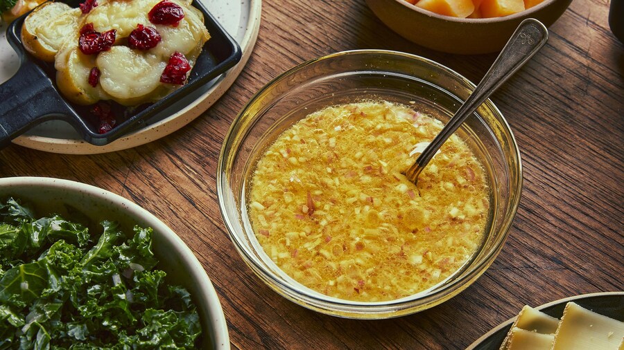 La marinade à raclette se trouve au centre d’une table, dans un bol en verre. Autour, il y a une assiette avec des patates gratinées et des canneberges cuites, un bol de kale, une assiette de fromage et un bol de rutabaga en cube.