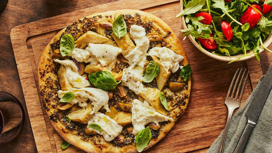 Sur une planche en bois, il y a une pizza aux artichauts et à la burrata et un bol de salade.
