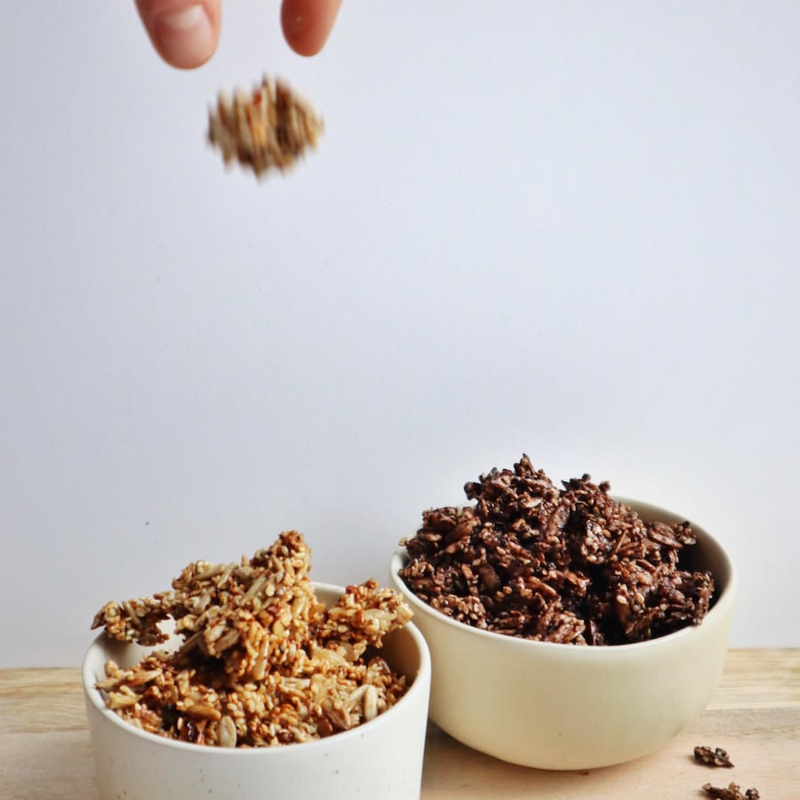 Sur une table, deux bols remplis de granolas préparés de deux façons, et une main laissant tomber un morceau de granola vers un des bols.